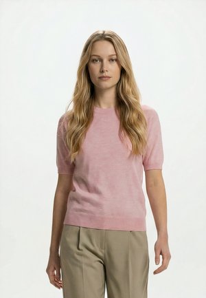 Donna con capelli biondi lunghi che indossa una maglietta rosa chiaro a maniche corte e pantaloni beige, in piedi davanti a uno sfondo bianco uniforme.