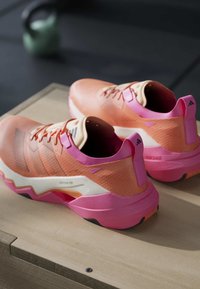 Paire de chaussures de sport roses et orange avec des semelles blanches reposant sur une surface en bois, équipement de gym flou en arrière-plan.