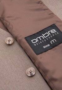 Tissu de veste marron avec deux boutons beige et une étiquette noire brodée en blanc indiquant « Ombre Quintessence Size M ».