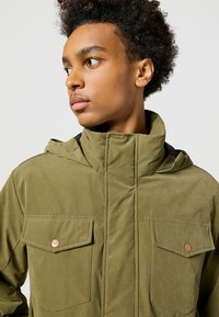 Jasnozielona kurtka z kapturem z dwoma kieszeniami na klapie, wysokim kołnierzem i gładką fakturą. Miedziane akcenty dekorują kieszenie.