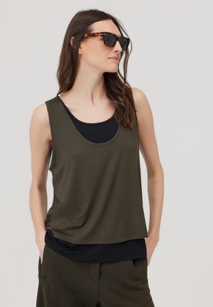 Kobieta nosząca ciemnozielony top bez rękawów na czarnym topie, pasujące ciemnozielone spodnie i okrągłe okulary przeciwsłoneczne w oprawie w kolorze żółwia, patrząca na bok.