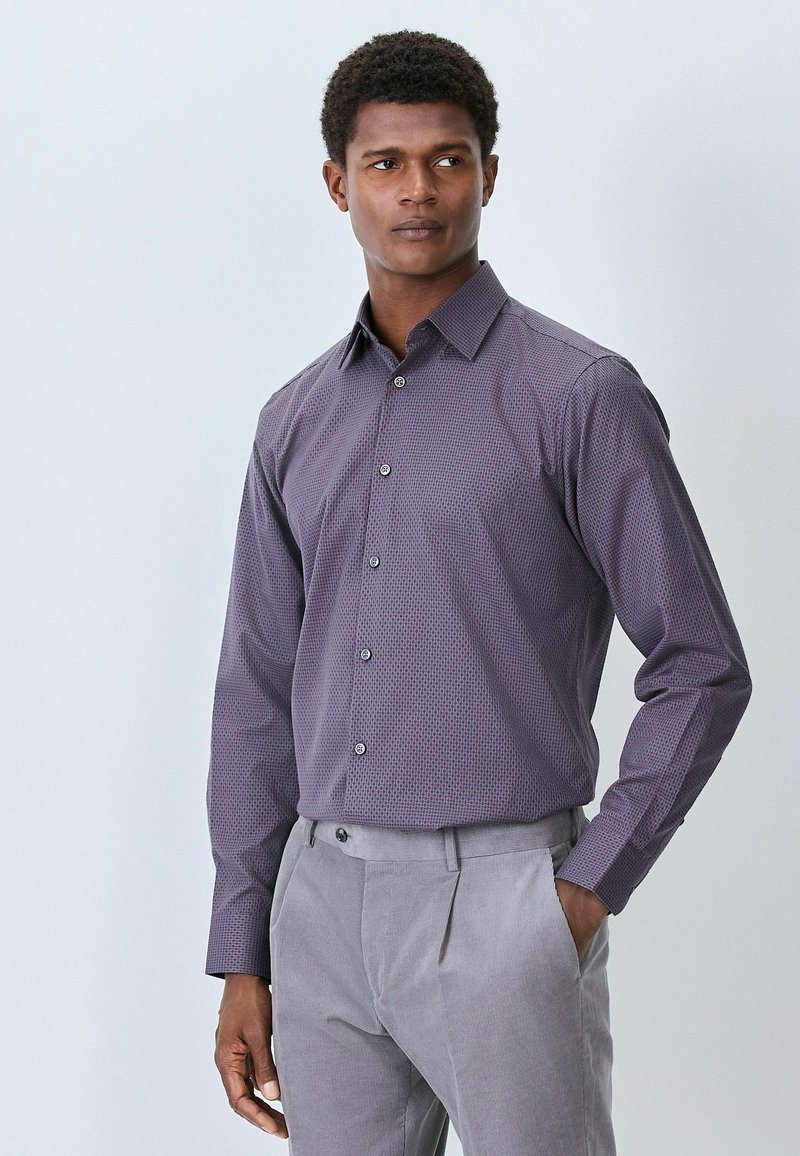 Herren Langarmhemd in Lila mit geometrischem Muster, kombiniert mit hellgrauen Hosen. Verfügt über einen Button-Down-Kragen und Knöpfe an der Vorderseite.