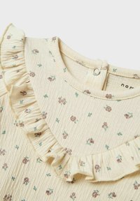 Blouse de couleur crème avec un petit imprimé floral, col froncé et fermeture à boutons à l'arrière. Tissu texturé avec une sensation légère et douce.