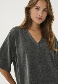 Young woman with long hair wearing a sparkly dark gray V-neck top, looking slightly to the side on a plain light background.