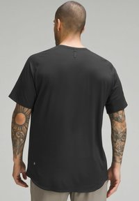 Chemise noire à manches courtes avec une texture, dotée d'une détail de couture au dos et d'un logo en bas de l'ourlet. Tatouages visibles sur le bras.
