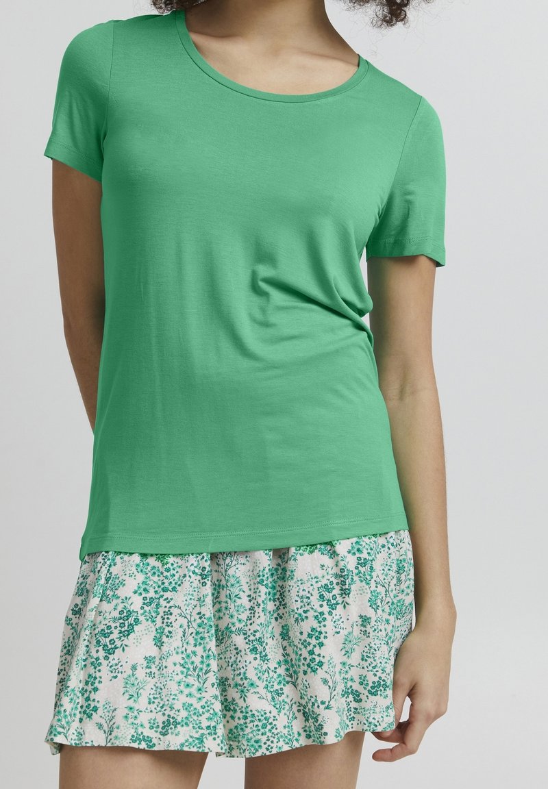 Grön t-shirt med kort ärm och rund halsringning, matchad med en vit kjol med gröna blommönster och lagerdesign.