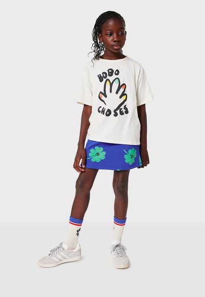 Vit t-shirt med svarta tryck och färgglad handdesign, kombinerad med en blå kjol med gröna klövermönster. Grå sneakers och randiga strumpor.