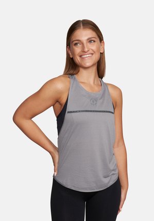 Uśmiechnięta kobieta o długich włosach, ubraną w szary bezrękawnik sportowy i czarne legginsy, stojąca z jedną ręką na biodrze.