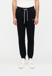 Μαύροι φόρμες με ελαστική μέση και ρυθμιζόμενο κορδόνι. Διαθέτουν λευκό κεντημένο λογότυπο στο αριστερό μπουτόνι. Φοριούνται με λευκά αθλητικά παπούτσια.