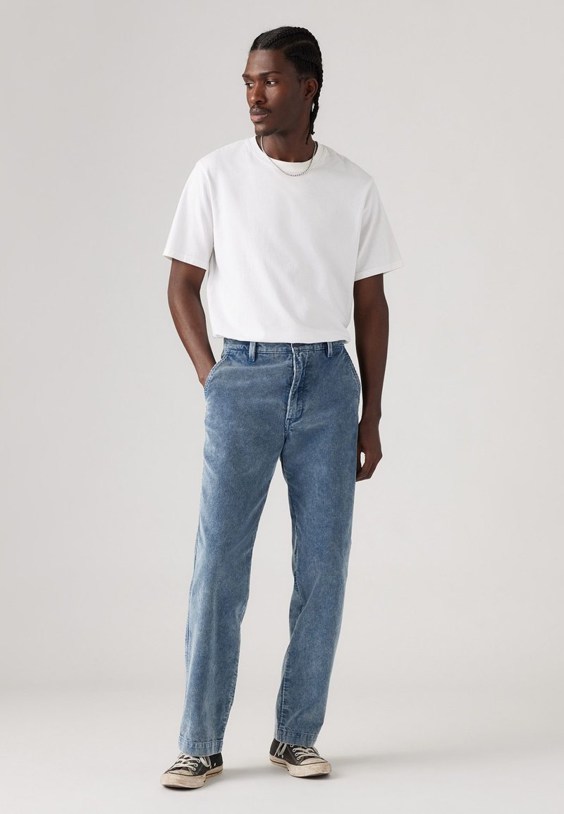Weißes Kurzarm-T-Shirt, hellblaue Jeans, lockere Passform, sichtbare Nähte, abgewetzte schwarze Sneakers mit weißen Sohlen, stehend mit den Händen in den Taschen.