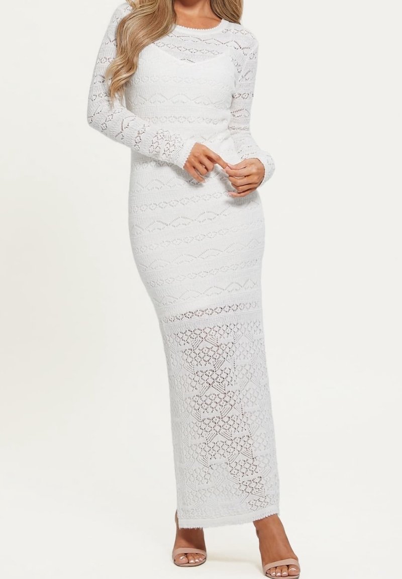 Longue robe blanche en dentelle avec motifs tricotés, manches longues et silhouette ajustée. Le tissu est transparent, révélant une texture subtile.