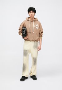 Ruda su gobtuvu džemperis su balta logotipu, kreminės kelnės su pilkais dėmėmis ir juodi sportbačiai. Modelis laiko juodą krepšį.