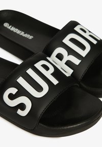 Ciabatte nere con ampia cinghia caratterizzate da lettere bianche in grassetto che spellano "SUPRDR." Suola strutturata, superficie della cinghia liscia e senza ulteriori decorazioni.