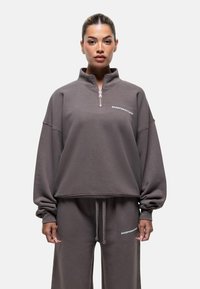 Felpa grigia con zip e colletto alto, fit ampio e testo ricamato. Tessuto morbido e maniche larghe. Abbinata a pantaloni sportivi coordinati.