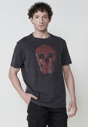 T-shirt en coton noir avec une finition usée, présentant un graphique de crâne rouge et du texte en bas. Manches courtes et col ras-du-cou.