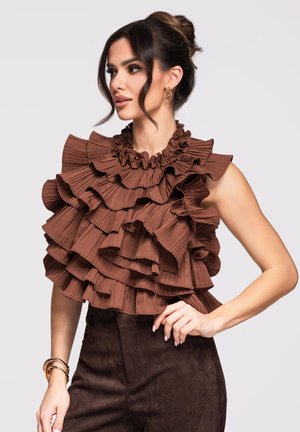 Kobieta o ciemnych włosach związanych w kok, ubrana w brązowy top bez rękawów z kilkoma warstwami falbanek i ciemnobrązowe spodnie, pozująca z jedną ręką na biodrze.