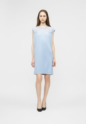 Jonge vrouw die staat, gekleed in een korte, mouwloze lichtblauwe jurk met een doorschijnend bovenpaneel, gecombineerd met zwarte hoge hakken, met een neutrale uitdrukking.