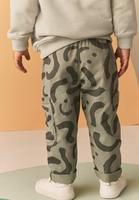 Grüne Cargo-Hosen mit schwarzen abstrakten Mustern, aus einem robusten Stoff, mit Seitentaschen und umgeschlagenen Säumen, kombiniert mit weißen Sneakers.