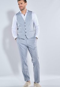 Hellgrauer dreiteiliger Anzug mit Weste, taillierter Hose und weißem Hemd. Enthält Knopfdetails und glatte Stofftextur. Helle Schuhe.