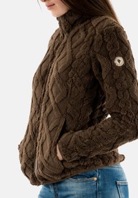 Giacchetto in pile marrone con un motivo a diamante testurizzato, colletto alto, tasche laterali e una patch del logo sulla manica sinistra.