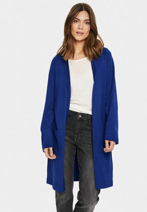 Blauw vest met lange mouwen, open voorkant, twee zakken en een relaxte fit, gedragen over een lichtgekleurd topje en donkere jeans.