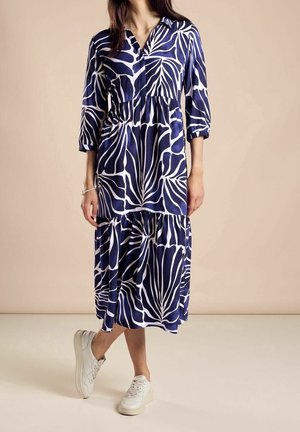 Marineblauwe jurk met een wit abstracte bladpatroon, kraagontwerp, driekwartmouwen en een gelaagde rok. Draagt met witte sneakers.