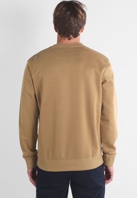 Sweatshirt khaki avec col rond, manches longues et poignets côtelés. Présente un petit détail d'étiquette sur l'épaule gauche. Texture lisse.