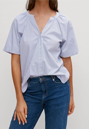 Kvinde iført en kortærmet, lyseblå og hvide stribet bluse med knapper, stoppet ned i mørkeblå jeans mod en ensfarvet baggrund.