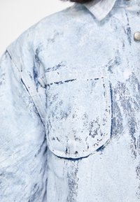Lys blå denimskjorte med et teksturert mønster, en lomme med klaff og knappestolpe. Stoffet har en falmet, slitt fremtoning.