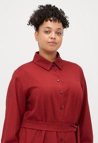 Vestido longo vermelho com colarinho, abertura frontal com botões e cinto na cintura. Apresenta uma textura suave e botões pretos, criando um design versátil.