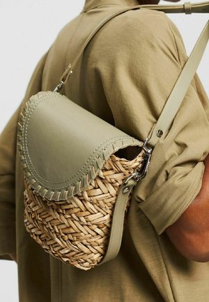 Persoon met een beige shirt met korte mouwen draagt een geweven rieten en leren schoudertas met gestikte klep over de schouder.