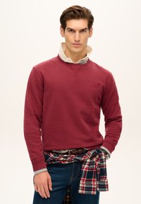 Marlot sweatshirt med rund hals og ribbede manchetter, lagd over en grå skjorte med krave. Parret med mørkeblå jeans og en ternet skjorte bundet omkring taljen.