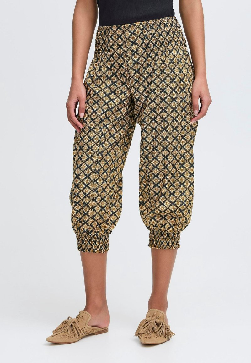 Femme portant un pantalon court avec un motif géométrique noir, doré et beige et des chaussures beiges à enfiler avec des pompons sur fond blanc.