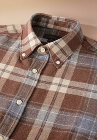 Camicia di flanella a quadri marrone e blu con colletto button-down e bottoni bianchi. Il tessuto ha un aspetto morbido e texturizzato.