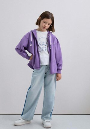 Sudadera con capucha y cremallera morada con cordones blancos, usada sobre una camiseta blanca con un diseño de calavera. Vaqueros de pierna ancha en azul claro con rayas laterales en azul.