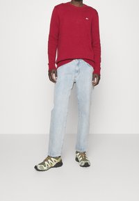 Czerwony sweter na drutach z prążkowanymi mankietami, jasnoniebieskie jeansy o prostych nogawkach oraz zielone sneakersy moro z czarnymi akcentami i beżowymi detalami.