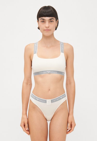 Cream-colored bra and thong set made of soft fabric with gray branded elastic straps. Features a square neckline and minimalistic design.