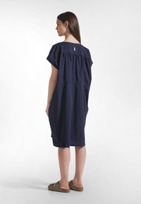 Marineblaues, kurzärmeliges Kleid mit gerafftem Rücken, knielang, aus weichem Stoff. Paar braune Sandalen mit Riemen, stehend auf hellem Hintergrund.