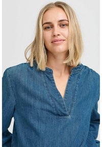 Camicia di denim blu con scollatura a V profonda, maniche lunghe e vestibilità rilassata. Il tessuto ha una consistenza morbida con una leggera lucentezza.