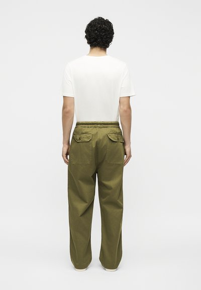 Άτομο με σγουρά μαλλιά που φοράει απλό λευκό μπλουζάκι και φαρδιά χακί παντελόνια, στέκεται γυρισμένο προς τα πίσω μπροστά σε λευκό φόντο.