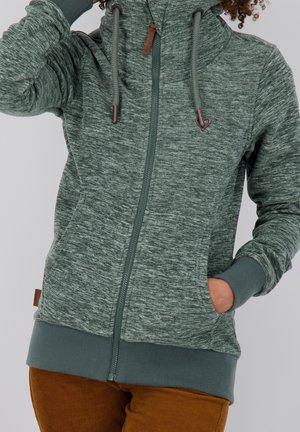 Personne portant un sweat à capuche vert à fermeture éclair avec cordons, main dans la poche, associée à un pantalon en velours côtelé marron, sur un fond uni.