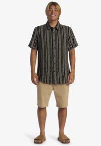 Camisa de manga corta blanca y negra con un estampado, pantalones cortos beige y sandalias beige. Tejido suave, corte relajado, con patrones de líneas horizontales y verticales.
