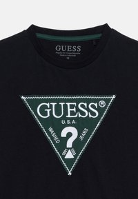 Μαύρο πουκάμισο με πράσινο τριγωνικό ύφασμα λογότυπο Guess που διαθέτει ένα λευκό ερωτηματικό και το κείμενο "GUESS U.S.A. Washed Jeans."
