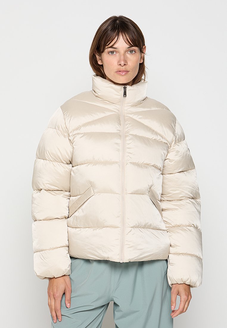 Kobieta w kremowej kurtce puchowej z zamkiem z przodu i jasnoniebieskich spodniach, stojąca na tle gładkiego, jasnego tła.