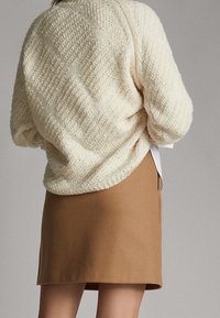 Le pull en laine crème présente un motif texturé et une coupe décontractée ; associé à une jupe mini ajustée en tissu lisse de couleur marron.