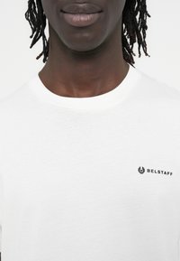 Balti medvilniniai marškinėliai su apvalia iškirpte ir mažu juodu „Belstaff“ logotipu kairėje krūtinės pusėje. Lygus audinys, kasdienio stiliaus dizainas.