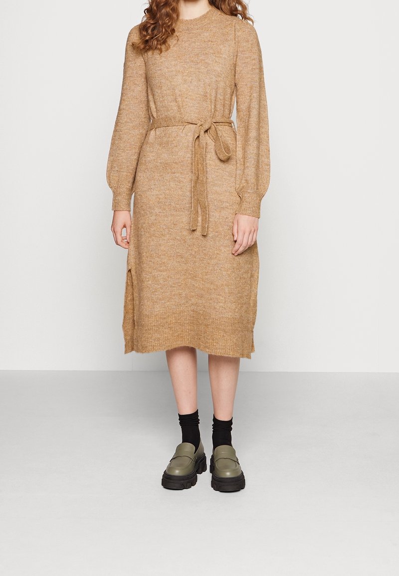 Femme portant une robe en maille camel mi-longue avec des manches longues et une ceinture nouée, associée à des chaussettes noires et des mocassins épais olive.