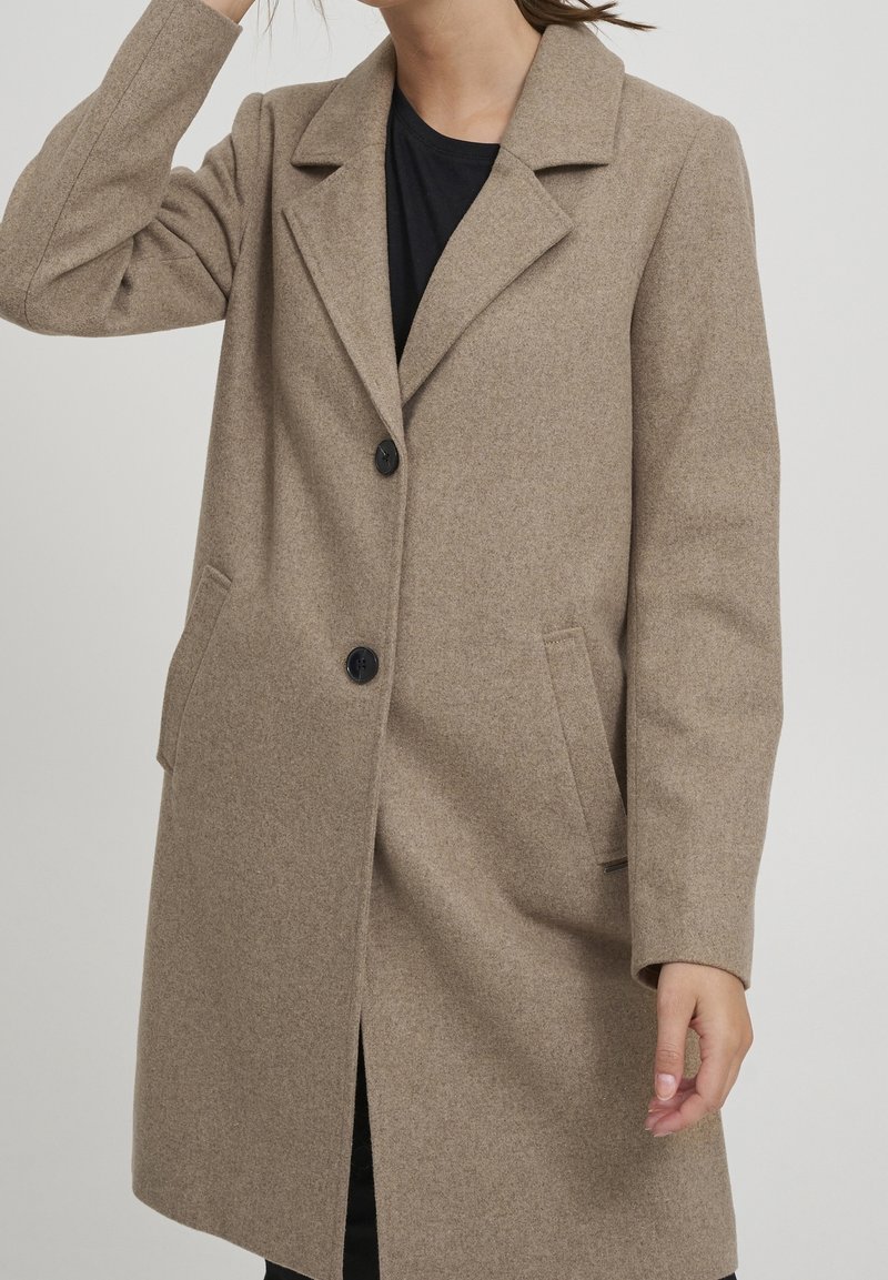 Manteau beige en mélange de laine avec un col cranté, deux poches avant et des boutons noirs. Présente une coupe droite et une fente centrale au dos.
