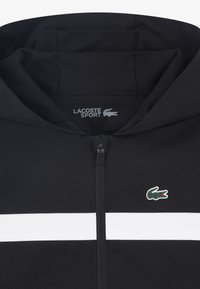 Μαύρο φούτερ με κουκούλα, με φερμουάρ μπροστά, λευκή οριζόντια ρίγα και πράσινη λογότυπο κροκόδειλου Lacoste στο αριστερό στήθος. Κατασκευάζεται από υφή ύφασμα.