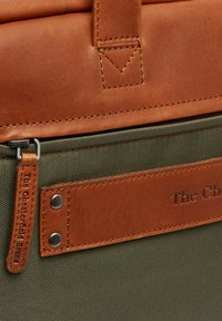 Brauner Leder-Obenklappe mit sichtbaren Nähten über olivgrünem Stoff. Enthält ein Lederschild mit eingraviertem Text und Metallelementen.